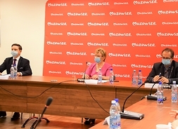Trzy osoby siedza za stołem w rzedzie, za nimi banner z logotypem samorządu Mazowsza.
