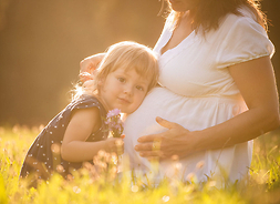 młoda kobieta w ciąży siedząca na słonecznej łące. Przytula się do niej mała dziewczynka