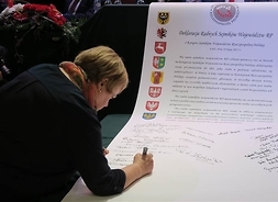 Radna podpisująca wielką płachte papieru z treścią deklaracji i godłami 16 województw