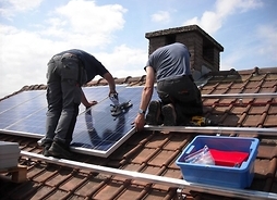 Dwóch robotników przyczepia panel do dachu, którego połowa już jest pokryta ogniwami