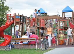 zdjęcie przedstawia plac zabaw z bawiącymi się dziećmi. Na ławkach siedzą opiekunowie i obserwują pociechy