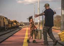 Chłopiec z plecakiem w kapeluszu biegnący na peronie od pociągu do czekającego na niego z rozpostartymi ramionami mężczyzny