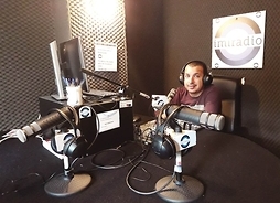 Studio radiowe z mikrofonami i komputerem, za stołem siedzi dziennikarz
