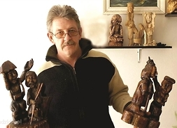 Zdjęcie rzeźbiarza w swetrze, okularach, z wąsami, trzymającego w rękach cztery małe drewniane rzeźby