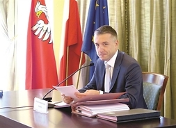 Samorządowiec w garniturze przy biurku, za nim flagi UE, Polski i województwa mazowieckiego