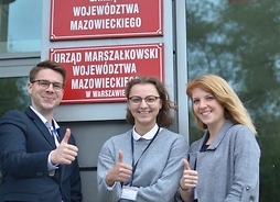 Troje studentów w eleganckich strojach pod tabliczką informującą, że tutaj mieści się Urząd Marszałkowski Województwa Mazowieckiego w Warszawie