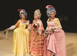 Kadr z przedstawienia w teatrze, na scenie trzy kobiety w wytwornych sukniach