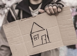 osoba bezdomna trzyma w ręce karton z nawysowanym flamasterem konturem domu