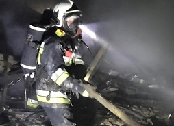 strażak w specjalistycznym ubraniu gasi pożar, obok zgliszcza domu