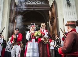 przed drzwiami kościoła stoi para nowożeńców w ludowych strojach w otoczeniu gości