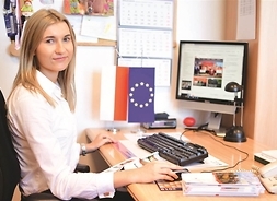 młoda dziewczyna pozuje do zdjęcia, siedzi przy biurku, na którym stoi komputer i leżą publikacje