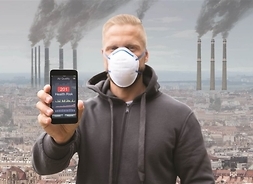 mężczyzna w masce na twarzy trzyma telefon z wyświetlonym na ekranie pomiarem PM 10 w tle dymiące kominy fabryki