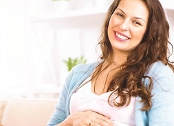 kobieta w zaawansowanej ciąży siedzi po turecku, trzyma się za brzuch i uśmiecha