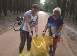 dwie kobiety na leśnej drodze trzymają worek w połowie wypełniony śmieciami