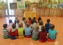 Dzieci siedzące na podłodze tyłem do patrzącego, słuchają prelekcji