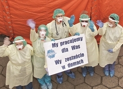 Pięcioro ubranych w kompletne kombinezony pracowników szpitala pozuje dozdjęciatrzymając przed sobą tablicę informacyjną z apelem do pacjentów.