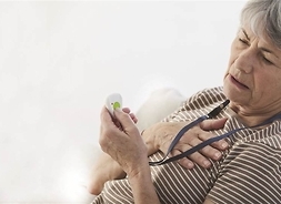 Seniorka siedzi oparta o poduszkę. Jedną dłoń trzyma na piersi, drugą wciska przycisk urządzenia alarmowego.