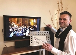 mężczyzna  batutą stoi pred telewizorem, na którym widać grającą orkiestrę