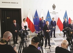 przy mikrofonach stoją mężczyźni w garniturach i kobiety w garsonkach, w tle mapy Polski u UE, z przodu stoją fotoreporterzy