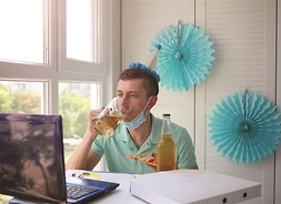 Młody mężczyzna siedzi przed ekranem komputera. W jednej ręce trzyma kufel z piwem, w drugiej kawałek pizzy. Maseczkę higieniczną ma zsuniętą pod brodę.