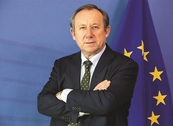 mężczyzna w garniturze pozuje do zdjęcia, widok do pasa, w tle flaga Unii Europejskiej