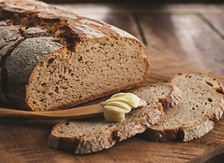 Bochenek chleba z odkrojonymi kilkoma kromkami. Na jednej leży drewniana szpatułka z masłem.