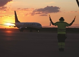 na płycie lotniska stoi samolot, na pierwszym planie mężczyzna kieruje ruchem, w tle zachód słońca