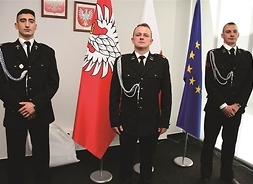 trzej strażacy w mundurach pozują do zdjęcia, pomiędzy nimi flagi Mazowsza i UE