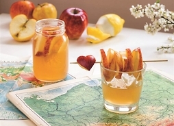 Szklanka soku jabłkowego, z leżącym na niej potyczkiem na który nabite są kawałki jabłka. Szlanka ustawiona na atlasie. W tle druga szklanka oraz jabłka.