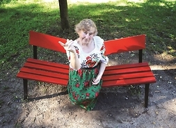 Starsza kobieta siedzi na ławce w parku. Ubrana jest w bluzkę i spódnicę z motywami ludowych haftów kwiatowych. Ma zawadiacką minę i wydaje się grozić nam palcem.