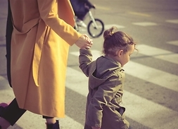 Kobieta w płaszczu trzyma za rękę kilkuletniego chłopca. Obydwoje przechodzą po pasach na przejściu dla pieszych