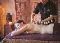 Kobieta leżyna brzuchu na stole do masażu, na odsłonięte plecy. Przy niej stoi masażysta w ciemnym stroju. Ustawia jej na plecach bańki chińskie.
