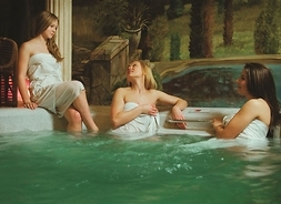 Trzy młode kobiety osłonięte tylko ręcznikami kąpielowymi rozmawiają z łaźni. Dwie stoją w wodzie, trzecia siedzi na krawędzi basenu.