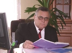 Mężczyzna w okularach, siedzący za biurkiem i z otwartą książką w dłoniach.