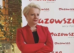 Kobieta stoi z założonymi rękami na tle banneru z napisem Mazowsze serce Polski