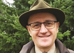 Uśmiechnięty mężczyzna w okularach i kapeluszu na głowie. Zdjęcie na tle drzew iglastych