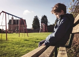 Chłopiec ze spuszczoną głową, siedzący samotnie na drewnianej ławce na placu zabaw