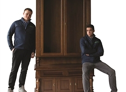 Dwóch młodych mężczyzn opartych o wysoki drewniany kredens