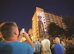 budynek z muralem przedstawiajacym pracownika w kasku, na pierwszym planie tłum ludzi, który jest skierowany w stronę budynku