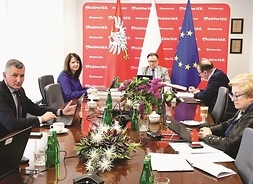 przy stole siedzi grupa osób, przed nimi monitory i kwiaty, w tle flagi Polski i Unii Europejskiej oraz ścianka z napisem Mazowsze