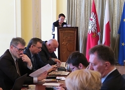 Przy stole siedzą radni klubu Prawa i Sprawiedliwości. W tle na mównicy przemawia radna Jolanta Koczorowska