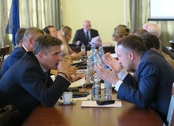 Radni Platformy Obywatelskiej siedzą przy stole, w tle przy stole prezydialnym siedzi wiceprzewodniczący Sejmiku Województwa Mazowieckiego Tomasz Kucharski