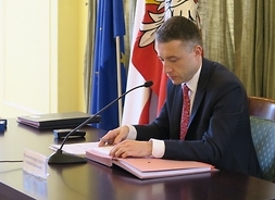 przewodniczący sejmiku siedzi przy stoliku, w tle flagi województwa, Polski i Unii Europejskiej