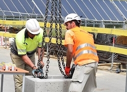 Dwóch robotników wiesza na łańcuchach betonowy blok z aktem erekcyjnym