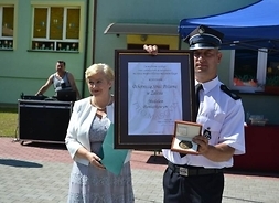 Dowódca jednostki prezentuje pamiątkowy dyplom i medal, obok przestawicielka samorządu