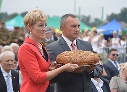 Z chlebem w dłoniach stoją starostowie dożynkowi, w tle uczestnicy święta
