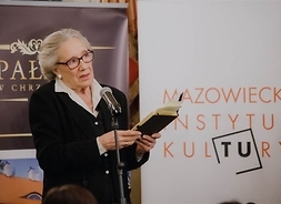 Aktorka z książką przy mikrofonie, w tle plakat Mazowieckiego Instytutu Kultury