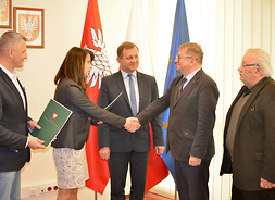 Uczestnicy podpisania umowy stoją na tle flag Mazowsza, Polski i Unii Europejskiej. Wicemarszałek Janina Ewa Orzełowska przekazuje podpisaną umowę.