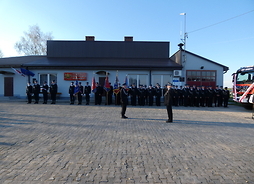 Przed remizą strażacką ochotnicy ze Szkop stoją w szeregu. Z lewej strony stoją poczty sztandarowe a z prawej nowe auto
