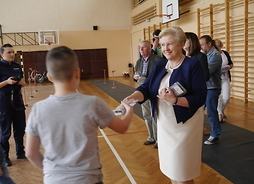 W centrum zdjęcia członek zarządu Elżbieta Lanc przekazuje zestaw uczniowi szkoły. W tle widać pozostałych uczestników i salę gimnastyczna.